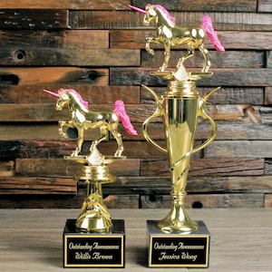 May include: Two gold-colored trophy cups with unicorn figurines on top. The unicorns have pink manes and tails. The base of the trophies are black and have engraved text that reads "Outstanding Awesomeness" followed by the name of the recipient. The text on the left trophy reads "Willie Brown" and the text on the right trophy reads "Jessica Wong".
