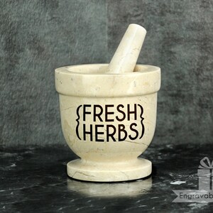 May include: A beige marble mortar and pestle set. The mortar has the words "FRESH HERBS" etched on the front. The pestle is inside the mortar. The set is on a black marble surface.