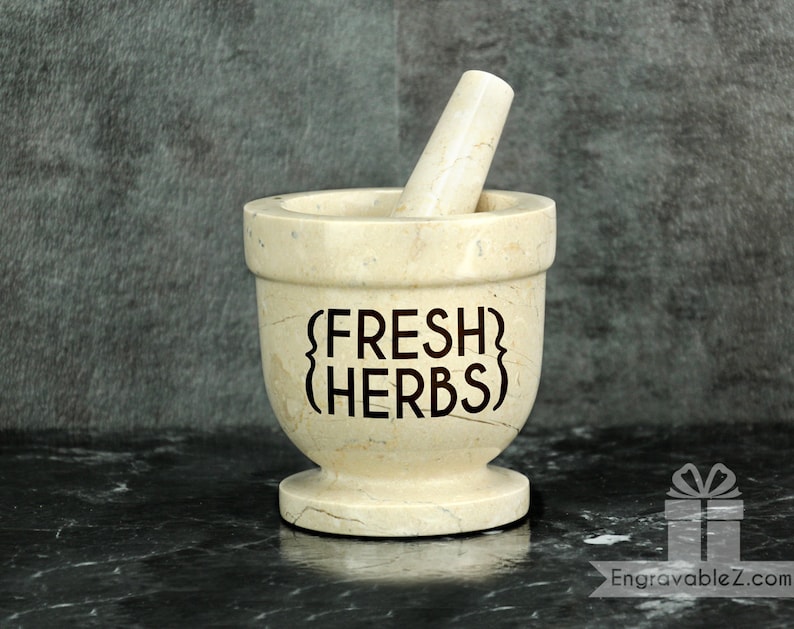 May include: A beige marble mortar and pestle set. The mortar has the words "FRESH HERBS" etched on the front. The pestle is inside the mortar. The set is on a black marble surface.
