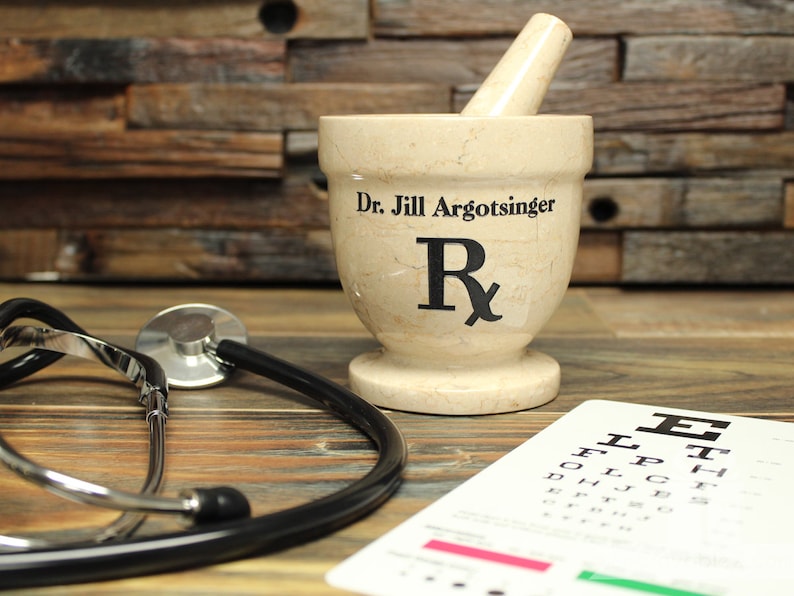 May include: A beige mortar and pestle with the text "Dr. Jill Argotsinger" and a prescription symbol. A stethoscope and an eye chart are in the foreground. The background is a wooden wall.