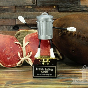 May include: A silver trash can figurine with black arms and white hands holding a gold trophy. The trophy has a black base with the words "Trash Talker Award" and "VICTOR GORDON" engraved on it. The figurine is standing on a wooden surface with a brown football and two leather football pads.