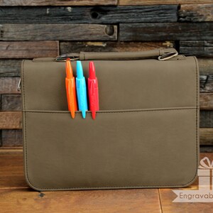 May include: A brown leather portfolio with a zipper closure and a pen holder. Three pens are inserted in the pen holder: one orange, one blue, and one red.