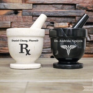 May include: Two personalised marble mortar and pestle sets. One is white with the text "Daniel Chung, PharmD" and an Rx symbol. The other is black with the text "Dr. Andrina Nguyen" and a caduceus symbol.