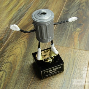 May include: A silver trash can shaped trophy with a black base. The trophy has a gold plaque that reads "Trash Talker Award" and "Victor Gordon".
