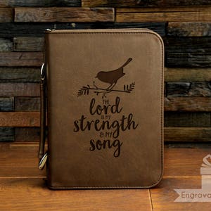 May include: A brown leather Bible cover with a zipper closure and a handle. The cover features an engraved design of a bird on a branch and the text "The Lord is my strength & my song."