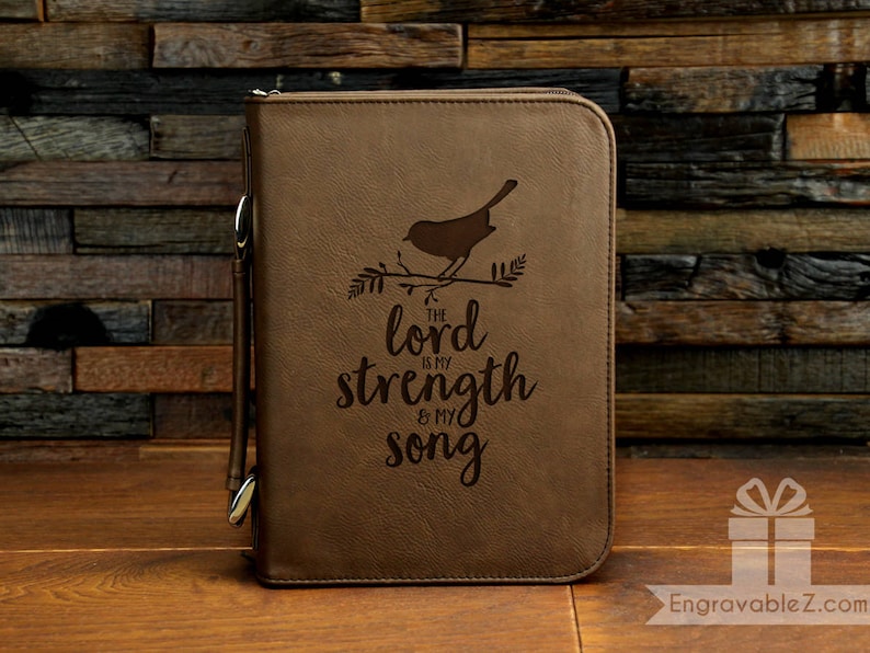 May include: A brown leather Bible cover with a zipper closure and a handle. The cover features an engraved design of a bird on a branch and the text "The Lord is my strength & my song."