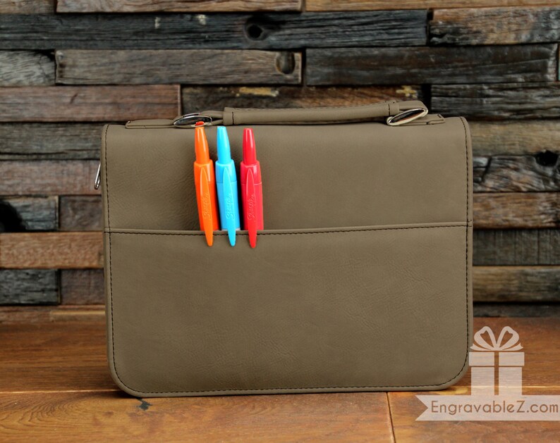 May include: A taupe-colored briefcase with a handle and a front pocket. Three pens in orange, blue, and red are in the pocket. The briefcase has a silver-colored clasp and the text "EngravableZ" is visible.