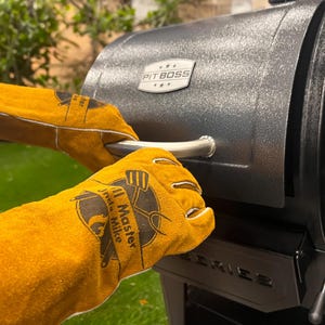 May include: Close-up of a black grill with a silver handle. The grill is being opened by a person wearing yellow leather gloves. The gloves have a logo that says "Grill Master Mike". The grill has the "Pit Boss" logo.