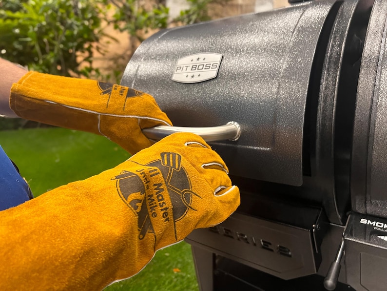 May include: Close-up of a black grill with a silver handle. The grill is being opened by a person wearing yellow leather gloves. The gloves have a logo that says "Grill Master Mike". The grill has the "Pit Boss" logo.