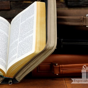 May include: A beige leather Bible cover with a zipper closure, open to reveal a yellowed Bible with pages of text. The cover is sitting on top of a stack of three brown leather cases.