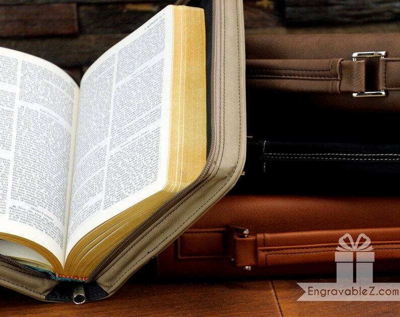 May include: An open Bible with yellowed pages is encased in a tan zippered cover. The cover is partially open, revealing the text within. Behind it, a stack of leather briefcases in shades of brown and black are visible.