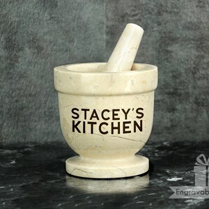 May include: A beige marble mortar and pestle set. The mortar features the text "STACEY'S KITCHEN". The pestle rests inside the mortar. The set is on a black surface.
