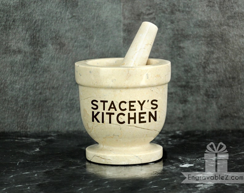 May include: A beige marble mortar and pestle set. The mortar features the text "STACEY'S KITCHEN". The pestle rests inside the mortar. The set is on a black surface.