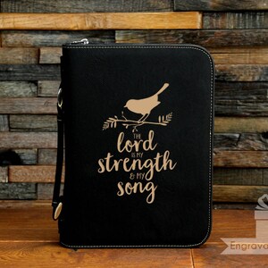 May include: Black zippered Bible cover with a gold bird and branch design. The text "The Lord is my strength & my song" is below the bird. The cover has a wrist strap and a decorative stitched border.