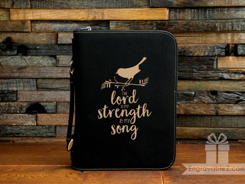 May include: Black zippered Bible cover with a gold bird and branch design. The text "The Lord is my strength & my song" is below the bird. The cover has a wrist strap and a decorative stitched border.
