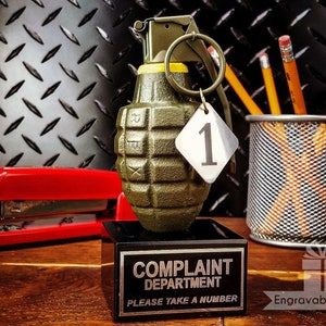 May include: A novelty desk accessory featuring a green grenade with a tag numbered "1". It sits on a black base with the words "COMPLAINT DEPARTMENT PLEASE TAKE A NUMBER". A red stapler and pencils in a metal holder are also visible.