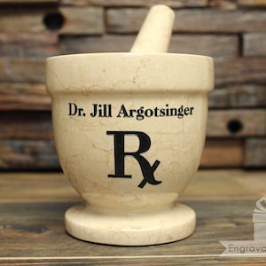May include: A beige mortar and pestle set with the text "Dr. Jill Argotsinger" and a black Rx symbol. The pestle rests inside the mortar. The mortar is on a wooden surface.