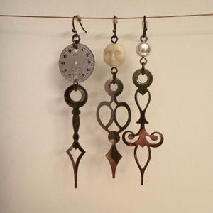 May include: Three unique steampunk-style earrings hanging from a wire. One features a clock face, another a face-shaped bead, and the third a pearl. Each earring has intricate metalwork in shades of bronze and silver, creating a vintage aesthetic.