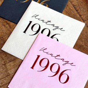 May include: Three square paper napkins in navy, white, and pink, each printed with the word "Vintage" and a year. The navy napkin has gold text for "1906", the white napkin has black text for "1906", and the pink napkin has red text for "1996".