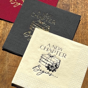 Puede incluir: Tres servilletas de papel cuadradas en rojo, antracita y crema. Cada servilleta tiene el texto dorado "A NEW CHAPTER" y una pila de libros atados con un lazo. La servilleta crema tiene el texto adicional "Begins..." y una rosa.