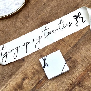May include: A white satin sash with the phrase "tying up my twenties" in black script. A small white square box tied with black string is also present. The items are on a wooden surface.