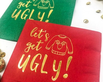 May include: Two square paper napkins, one green and one red, each featuring gold foil text that reads "let's get UGLY!" alongside a gold line drawing of a sweater with a reindeer graphic on the front, arranged on a white surface.