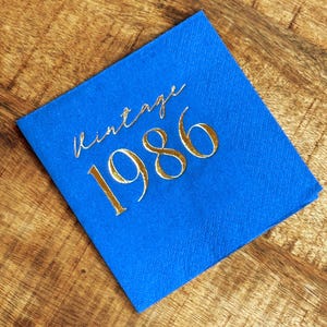 blue paper napkin with the gold text "vintage 1986". The napkin is made of a textured paper material and is set on a wooden surface. The text is in a cursive font.