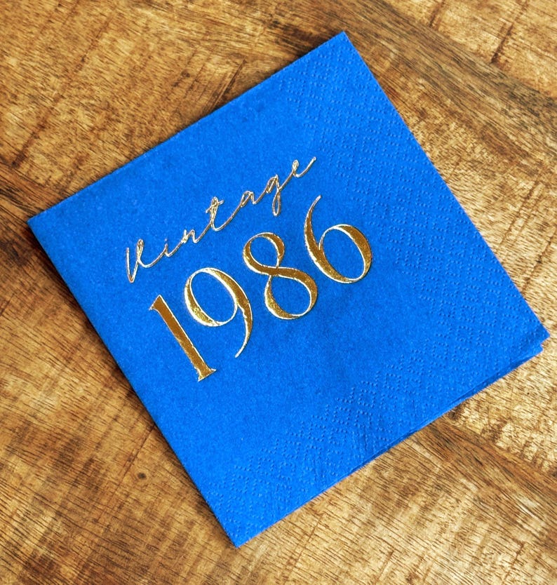blue paper napkin with the gold text "vintage 1986". The napkin is made of a textured paper material and is set on a wooden surface. The text is in a cursive font.