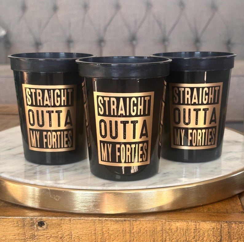 Three black stadium cups with gold printed text reading Straight Outta My Forties stand on a marble and gold tray.