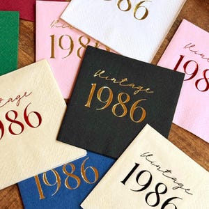 A collection of square paper napkins in various colors, including white, pink, and black. Each napkin is printed with the words "Vintage 1986" in gold or red metallic lettering, suitable for a birthday celebration or event.