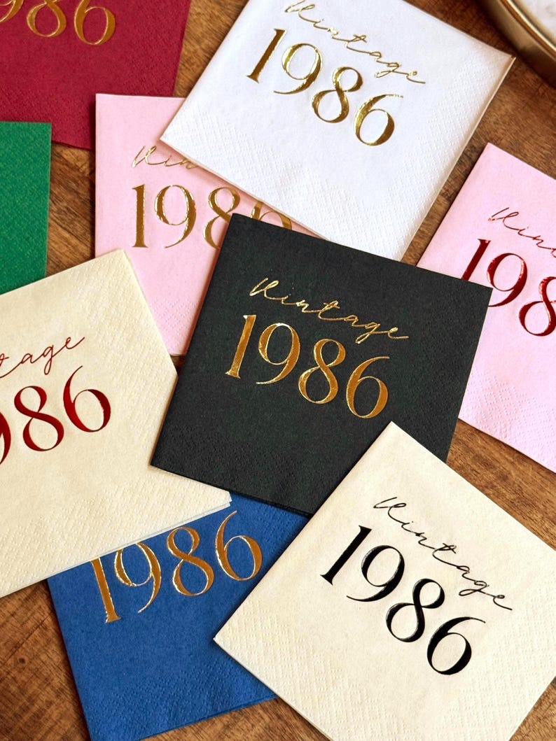A collection of square paper napkins in various colors, including white, pink, and black. Each napkin is printed with the words "Vintage 1986" in gold or red metallic lettering, suitable for a birthday celebration or event.