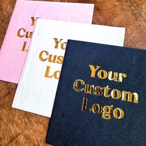 Könnte beinhalten: Drei Servietten mit individuellem Logo in Rosa, Weiß und Marineblau. Die Servietten sind quadratisch und mit goldfarbenen Buchstaben versehen, die "Your Custom Logo" lauten. Die Servietten liegen auf einer Holzoberfläche.
