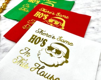 May include: Three white paper napkins with gold foil lettering and a cartoon image of Santa Claus wearing sunglasses. The text on the napkins reads "There's Some HO'S In This House".