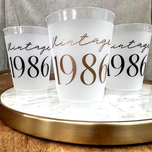 Three frosted plastic cups with "vintage" in script and "1986" in block letters, two with black text and one with gold text, sit on a white marble tray with a gold rim.