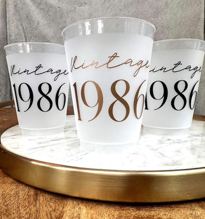 Three frosted plastic cups with "vintage" in script and "1986" in block letters, two with black text and one with gold text, sit on a white marble tray with a gold rim.