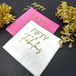 May include: White paper napkins with gold foil lettering that reads "Fifty & Fabulous".