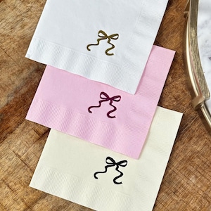 May include: Three square napkins with a bow design. The napkins are white, pink, and cream. The bows are gold, purple, and black.