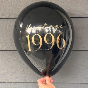 May include: A black balloon with gold text reading "Vintage 1996". The balloon is held by a hand. The balloon reflects the surrounding environment.