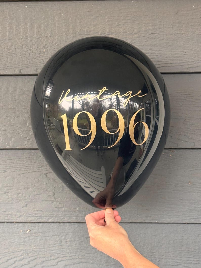 May include: A black balloon with gold text reading "Vintage 1996". The balloon is held by a hand. The balloon reflects the surrounding environment.