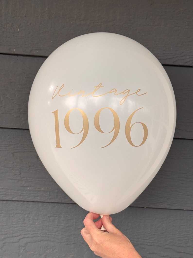 May include: A white balloon with gold text reading "Vintage 1996". The balloon is held by a hand. The background is a dark grey colour. The balloon is round and inflated.