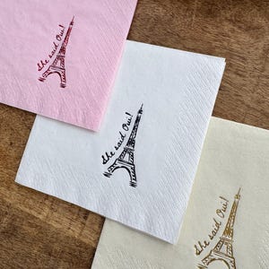 May include: Three paper napkins in pink, white, and cream, each printed with an Eiffel Tower and the phrase "She said Oui!". The napkins are displayed on a wooden surface, ideal for a wedding or engagement celebration.