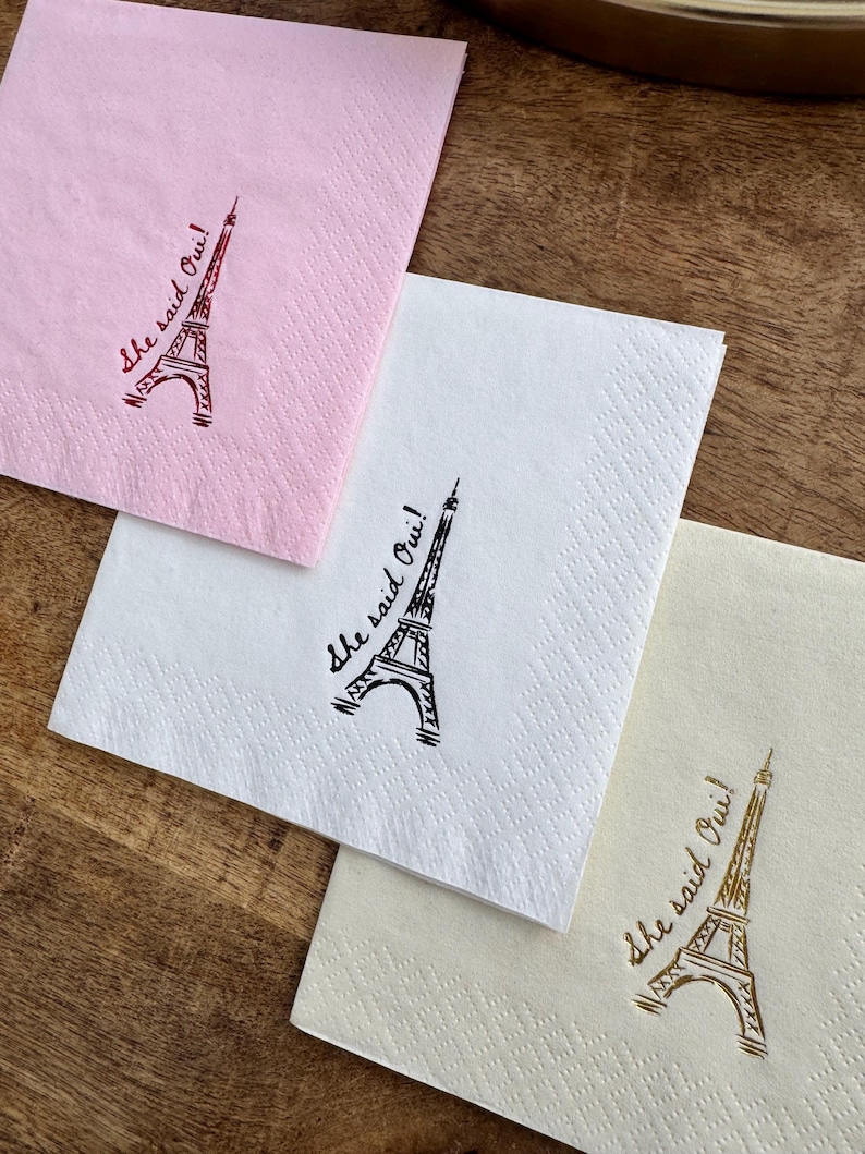 May include: Three paper napkins in pink, white, and cream, each printed with an Eiffel Tower and the phrase "She said Oui!". The napkins are displayed on a wooden surface, ideal for a wedding or engagement celebration.