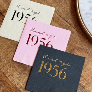May include: Three square paper napkins in cream, pink, and black, each with the word "Vintage" in cursive and the number "1956" in a bold font. The napkins are arranged on a wooden surface.