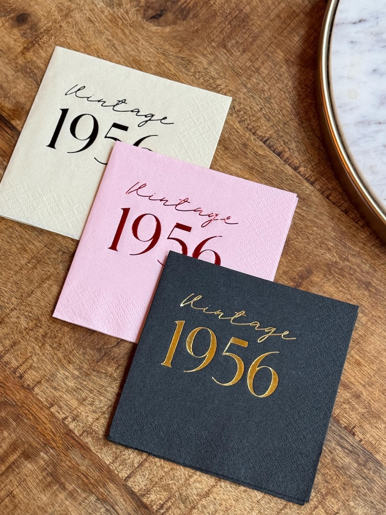 May include: Three square paper napkins in cream, pink, and black, each with the word "Vintage" in cursive and the number "1956" in a bold font. The napkins are arranged on a wooden surface.
