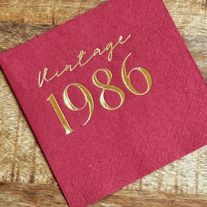 burgundy napkin with the gold text "vintage 1986". The napkin is made of a textured material and is set on a wooden surface. The gold text is in a cursive font.