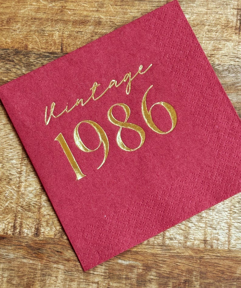 burgundy napkin with the gold text "vintage 1986". The napkin is made of a textured material and is set on a wooden surface. The gold text is in a cursive font.