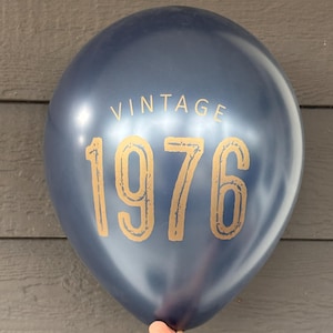 Puede incluir: Un globo azul marino con la palabra "VINTAGE" encima de los n&uacute;meros dorados "1976". El globo est&aacute; sostenido por una mano sobre un fondo de madera gris. Una decoraci&oacute;n festiva para un cumplea&ntilde;os.