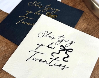 May include: Three white napkins with black ink and gold foil accents. The napkins feature the text "She's tying up her twenties" with a gold bow graphic.