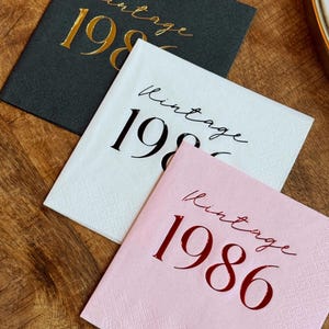Three square paper napkins are arranged on a dark wooden surface: a dark gray napkin with gold foil text, a white napkin with black foil text, and a pink napkin with red foil text, all reading "Vintage 1986."