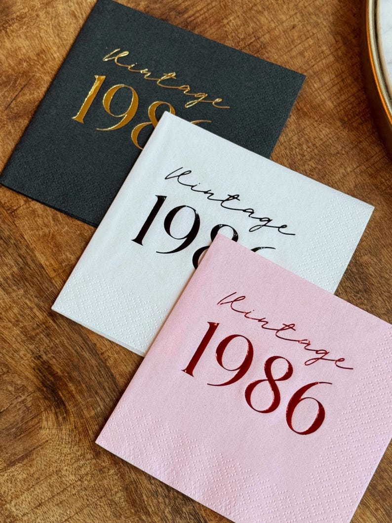Three square paper napkins are arranged on a dark wooden surface: a dark gray napkin with gold foil text, a white napkin with black foil text, and a pink napkin with red foil text, all reading "Vintage 1986."